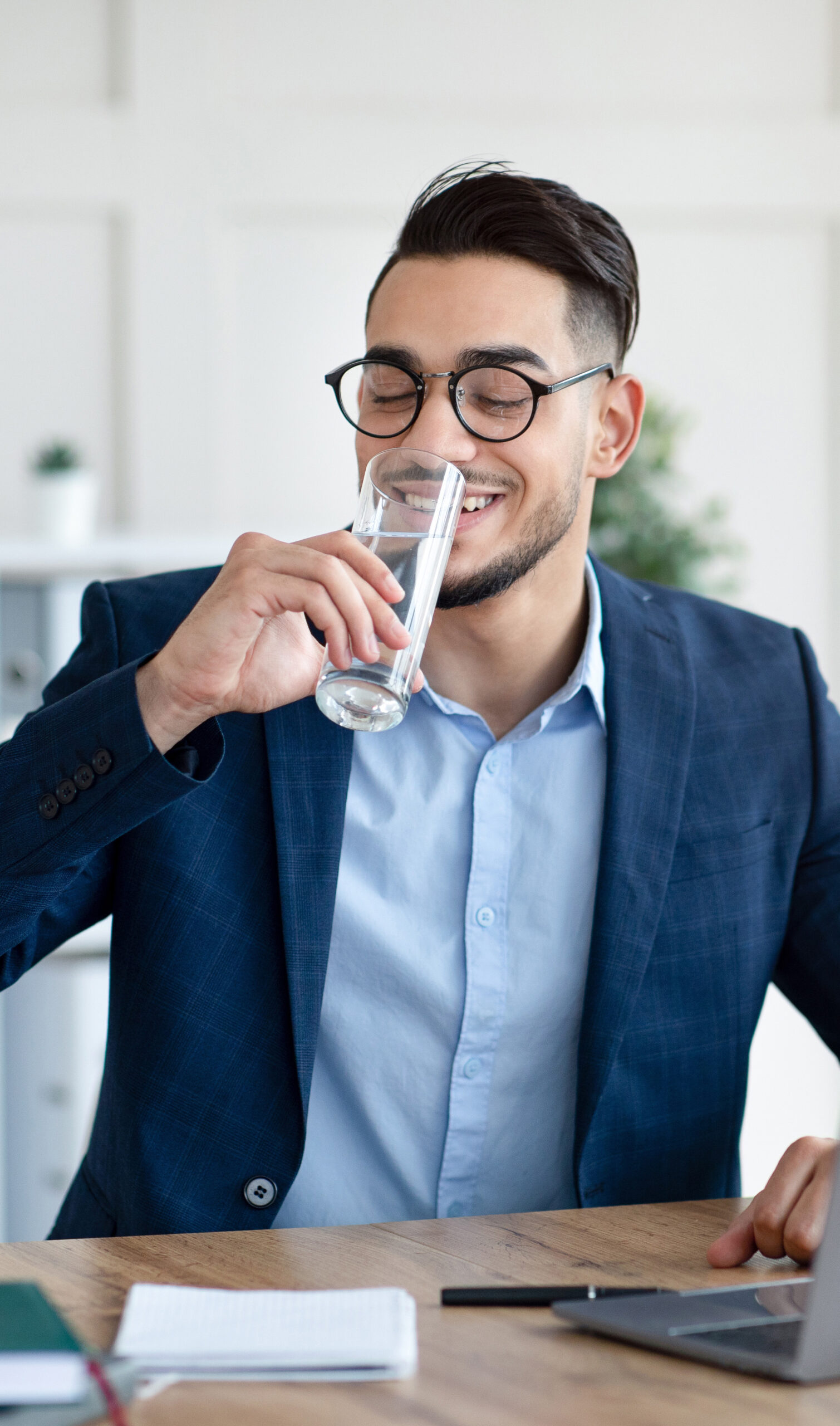 Cool Arab businessman in suit drinking water while working on laptop at modern office